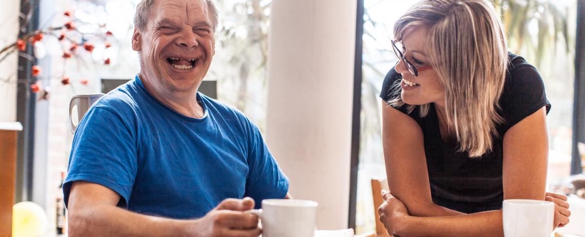 Man met verstandelijke beperking lachend aan tafel met begeleider